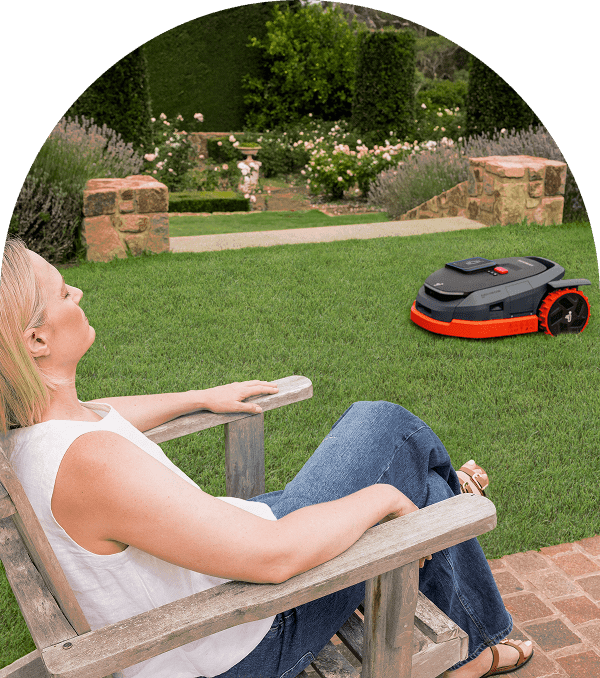 A woman relaxes outdoors while a robotic lawn mower works on the grass.